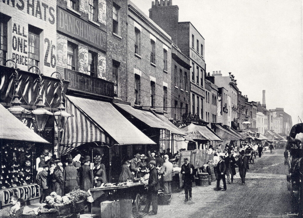 Black and white photograph from 1896 showing shops with awnings and barrows in front and customers,  to the right a horse and carriage