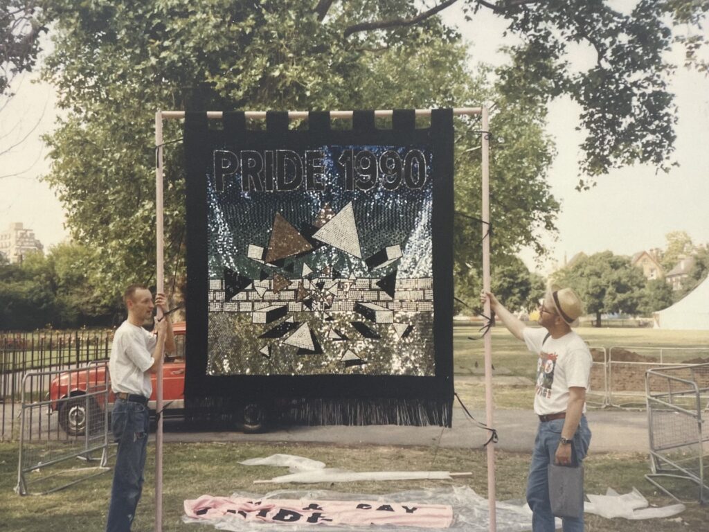 Archive photo of 2 men in a park holding up a large square banner with the words Pride 1990 on it